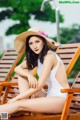 A woman in a white bathing suit and a straw hat sitting on a wooden chair.